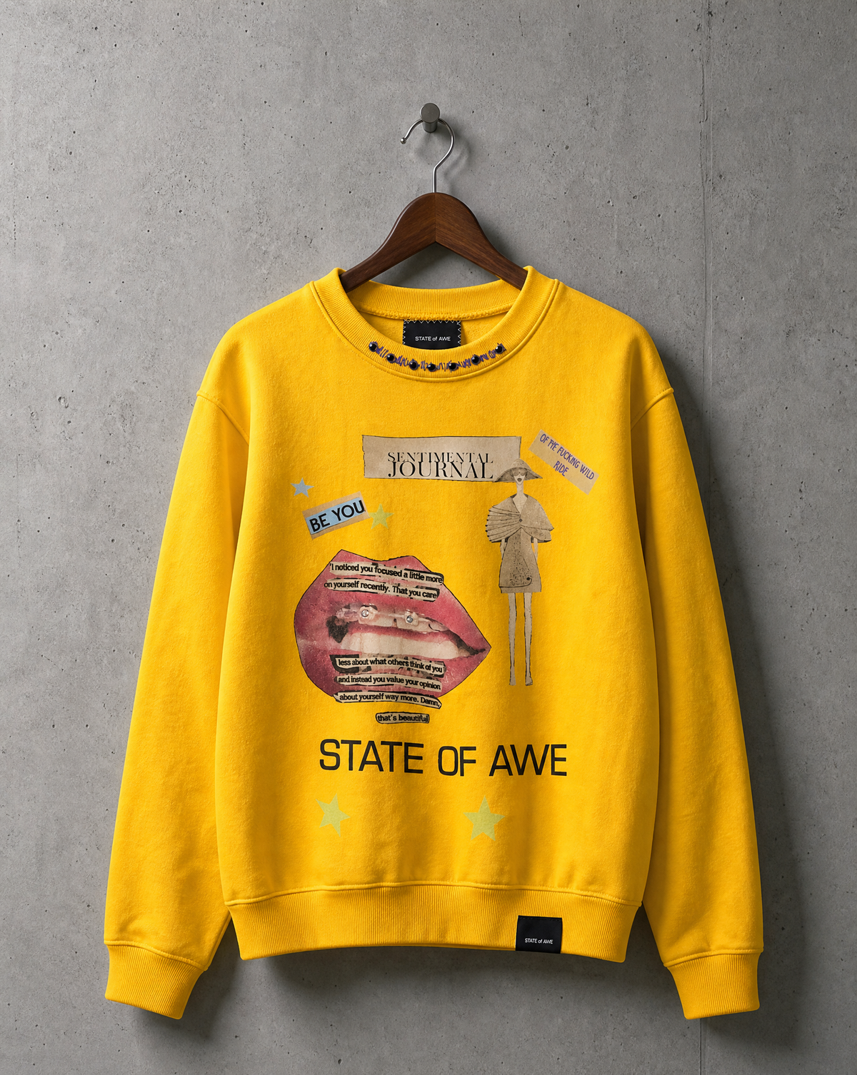 A bright yellow crewneck sweatshirt, one of a kind, hanging on a wooden hanger against a textured gray concrete wall. Features a graphic collage print with a pink lips illustration, a small figure, and text reading 'STATE OF AWE' and 'Sentimental Journey.'